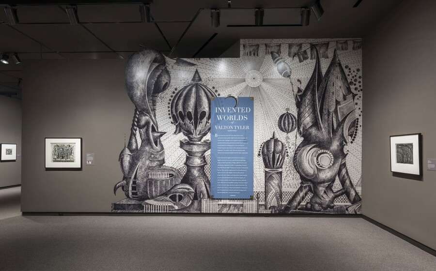 Exhibition title wall featuring an enlarged reproduction of a fantastical black-and-white drawing with introductory text on top of it. To the left and right are framed artworks hanging on taupe walls.