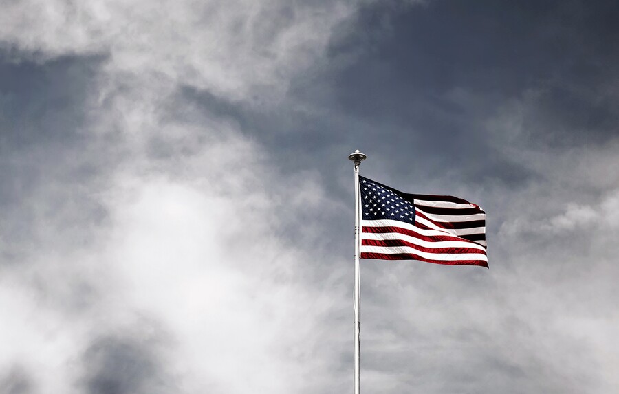 An American flag flies on a flagpole against white clouds and blue sky.