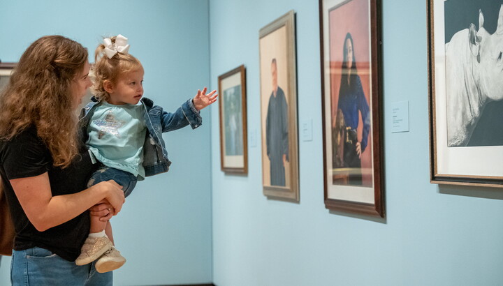 An adult holds a small child in her arms as the child gestures toward an artwork on the wall.
