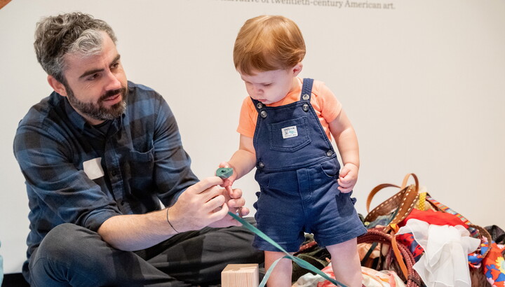 An adult sits on the floor next to a toddler as they both play with several props.