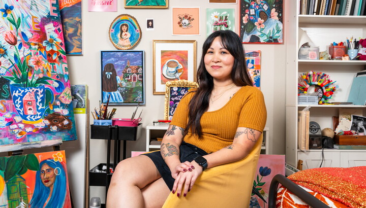 A smiling Asian woman with long dark hair is seated in a room with walls covered with colorful artwork.