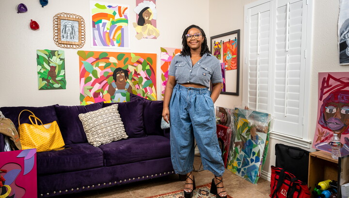 A smiling Black woman wearing glasses stands in front of a purple couch above which hangs many colorful artworks.
