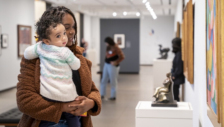 A Black woman holding a baby in her arms looks at a sculpture in a Carter gallery.