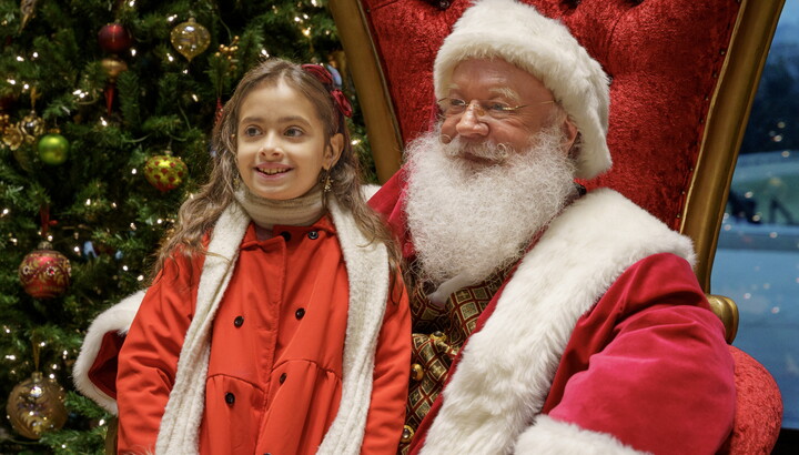 A young, light-skinned girl leans into Santa Claus who is sitting on his throne at the Carter.