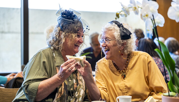 Two older White women, both wearing hats, laugh together as they enjoy sandwiches.