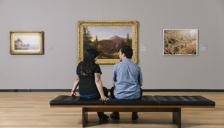 Two adults sit next to each other on a bench in a Carter gallery looking at art.