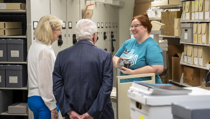 A smiling young White woman with red hair shows an object to a couple standing in a room lined with shelves..