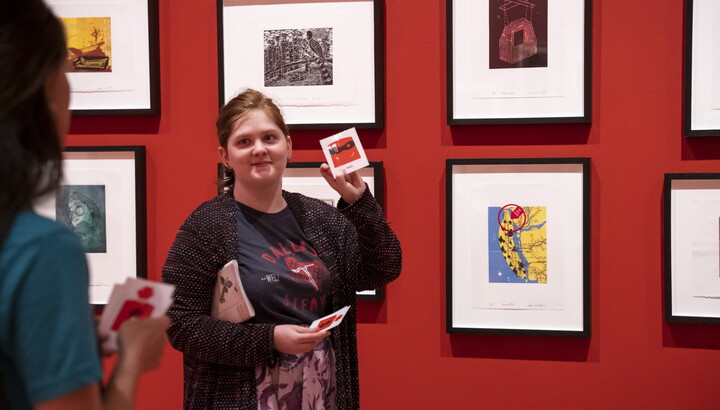 A young White woman stands in front of artwork in a Carter gallery holding up flash cards for someone out of the frame.
