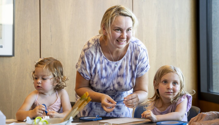 A White woman leans over a table in the Carter's Lounge where two children are making artwork.