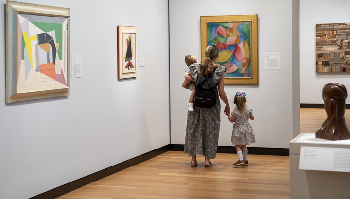 A woman and two children, one she holds on her hip and she hold the hand of the other, look at colorful art in a Carter gallery.