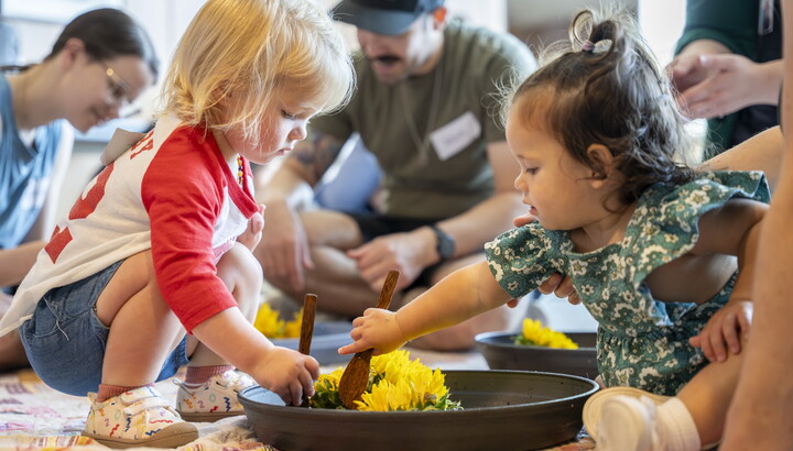 Two very young children play together with colored flowers in a bowl on the floor of a Carter gallery.