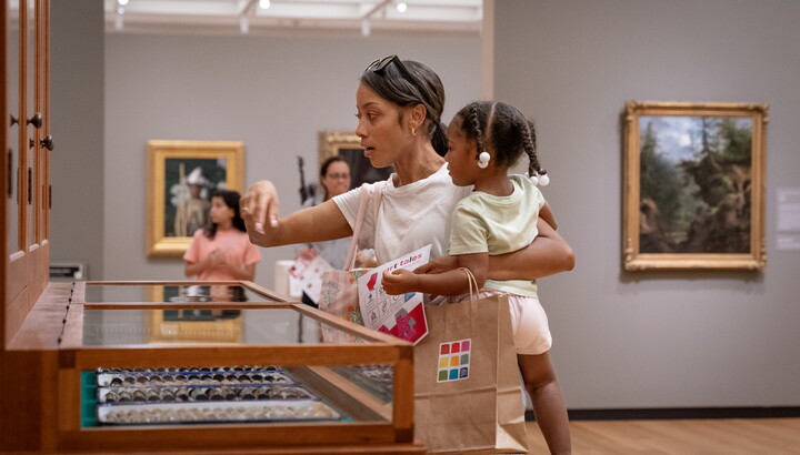 A dark-skinned woman holding a child on her hip points to some of the objects in "The Texas Cabinet."