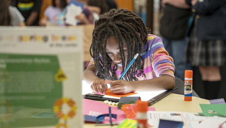 A black tween, focused on making art, leans over a table in the Carter's Library.