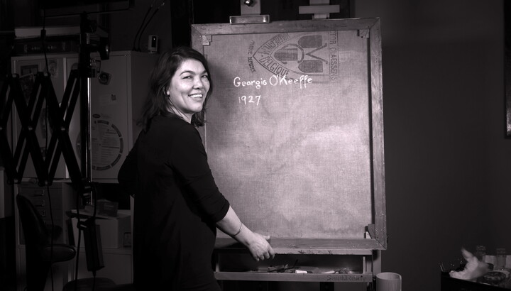 A black-and-white photo of a medium-skinned woman standing next to a canvas showing Georgia O'Keeffe's signature on the back.