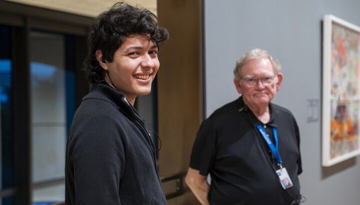 A smiling young, medium-skinned man with dark curly hair and an older White man with glasses, both in black shirts, stand in a Carter gallery.