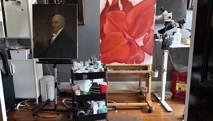 A painting of a red flower sits on an easel in a studio filled with other paintings.