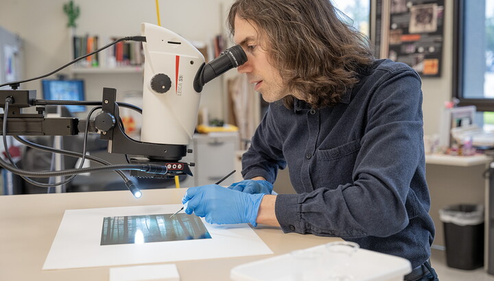 A White man with longish dark hair looks into a microscope-like device to view details on the photograph beneath it.