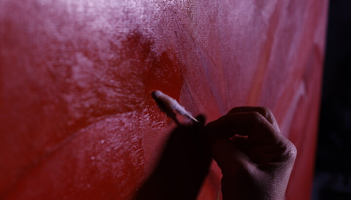 A close-up of hand holding a cotton swab with a dirty head next to a red painting. 