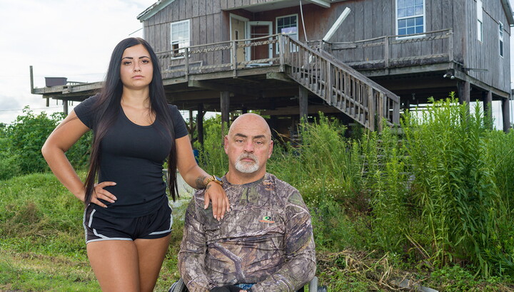 Two people outside of a house on stilts: a young woman with long dark hair stands next to a middle-aged man seated in a wheelchair.