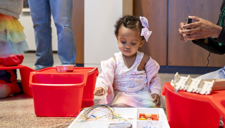A young Black child with a giant pink bow in her hair plays with art supplies on the floor of a Carter gallery.