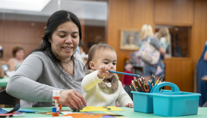 A medium-skinned woman with a baby on her lap sits at a table in the Carter's Library making art.