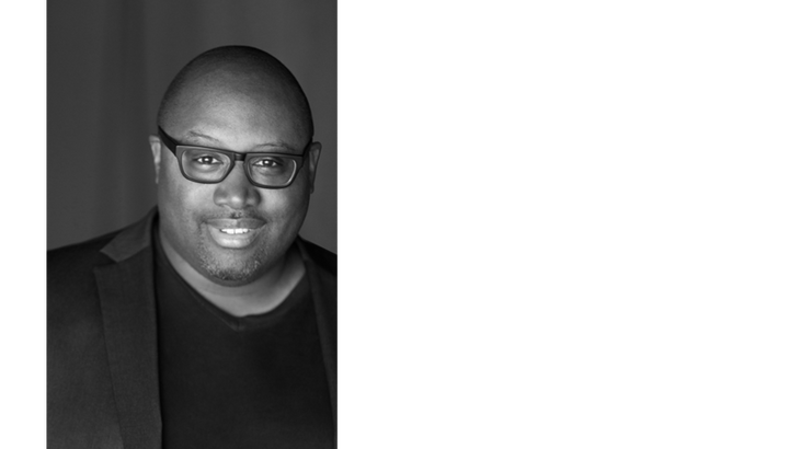 A black-and-white headshot of a bald Black man wearing glasses.