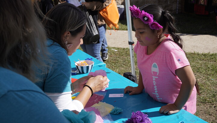 A woman leans over a table to show a medium-skinned child how to make an artwork.
