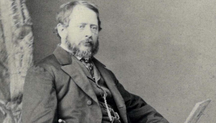 A black-and-white portrait photograph of a seated middle-aged White man with a beard and mustache wearing a three-piece suit, a cane resting by his side.