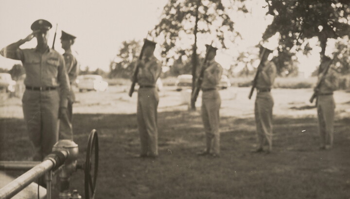 A slightly out-of-focus black-and-white photograph of a line of people in military uniforms, each holding a rifle, as one soldier salutes; tall trees and cars are in the background.