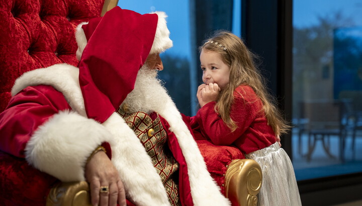 A young White girl leans on her elbow toward Santa who is seated in his throne.
