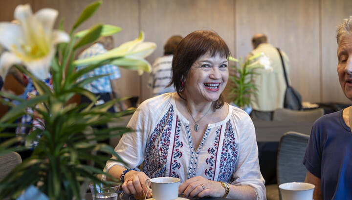 A smiling older White woman looks from behind a vase of flowers while seated at a table drinking tea.
