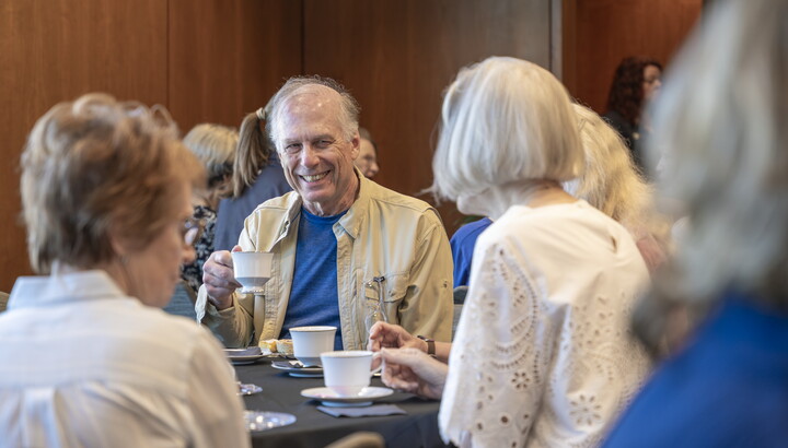 A smiling older White man drinks tea at a table with other guests.