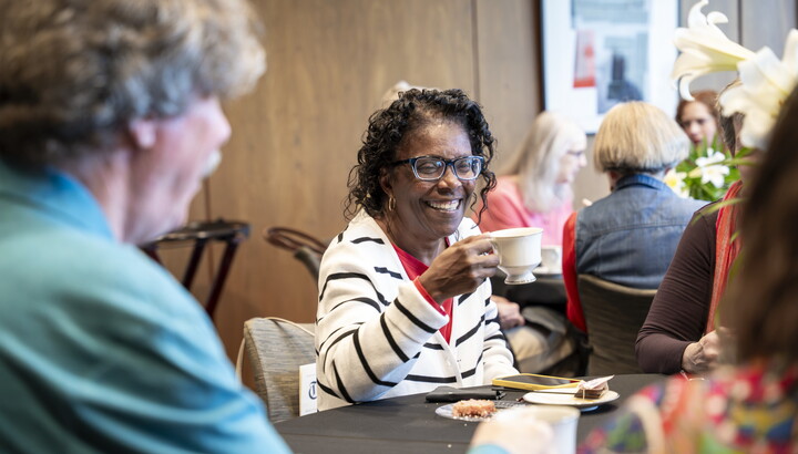 A smiling older Black woman holds up a cup of tea at a table in the Carter's Lounge.