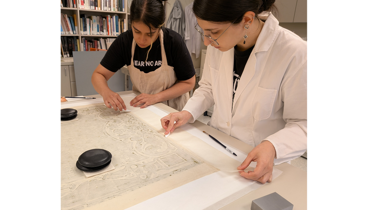 Two White women in lab coats work on an artwork laying on a table.