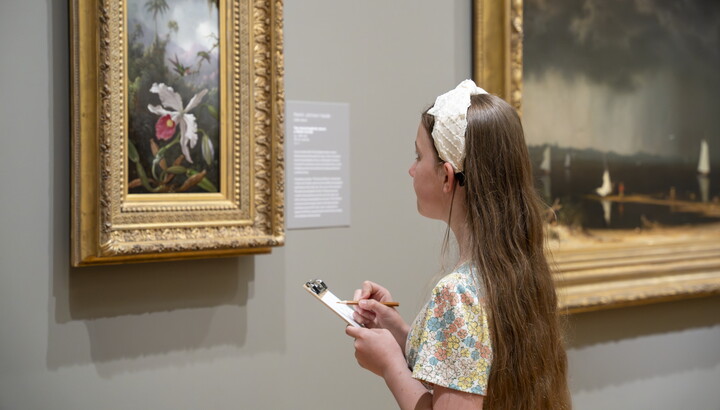 A White girl with long hair looks at an artwork in a Carter gallery and sketches it on a pad of paper.