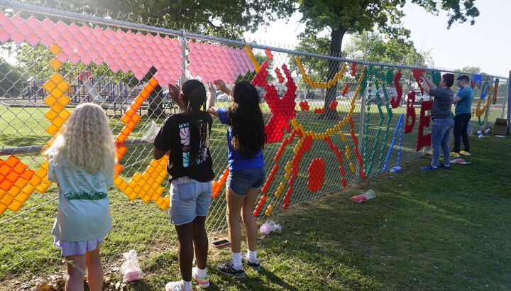 Children and adults use colored plastic pieces to decorate a chain link fence.