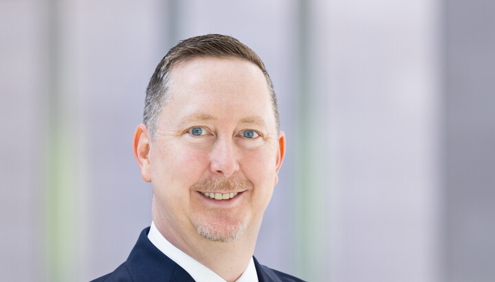 A headshot of a smiling White man with buzz-cut hair and goatee, wearing a blue suit, arms crossed.