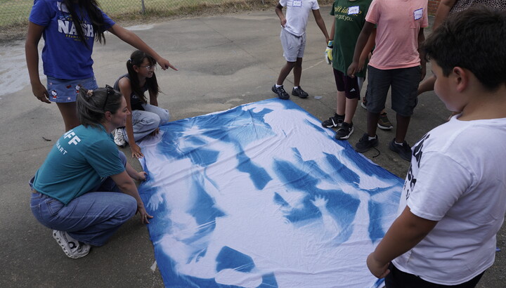 A group of middle-school-aged children and an adult lay out a large piece of fabric with blue silhouettes.