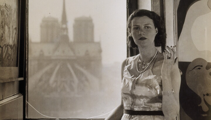 A black-and-white photograph of a young White woman standing in front of a window through which you can see the back of a church.