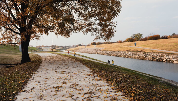 A color photograph of a late fall landscape with a river disappearing into the distance and people running on well-groomed paths.