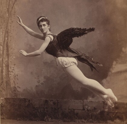 Black-and-white photograph from the 1880s of a woman wearing a dark corset with black feathered wings attached to the back and posed as if she is flying