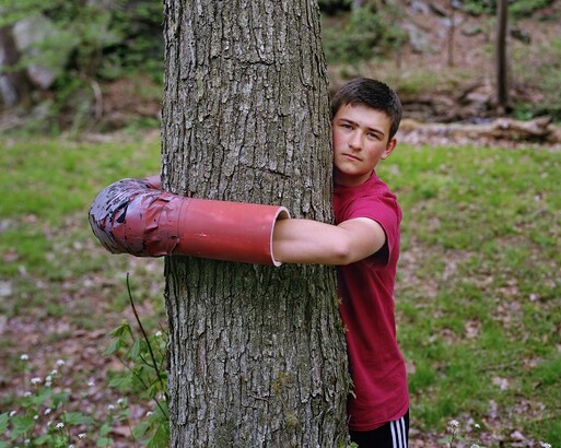 Boy with arms around a tree; his hand and forearms are inside a tube allowing him to wrap completely around the tree