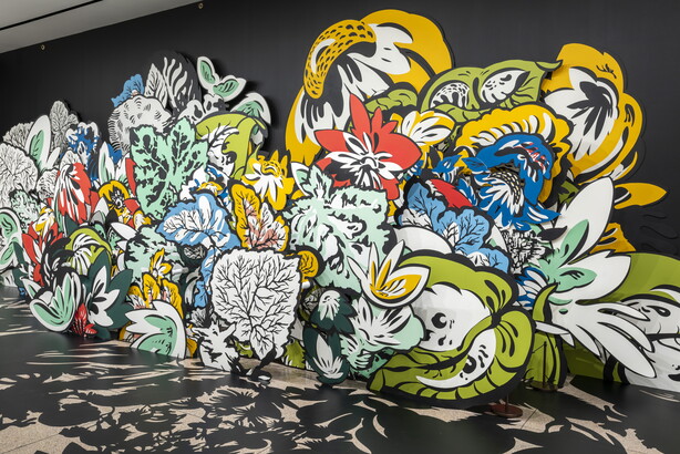 An installation photo of large wood cut-outs of flowers and leaves, some brightly-colored, against a black wall with shadows projected on the floor in a Carter gallery. 