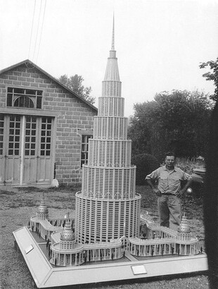 A black-and-white photograph of a White man standing behind an elaborate model of a building with seven tiers, like a cake, surrounded by a columned portico.