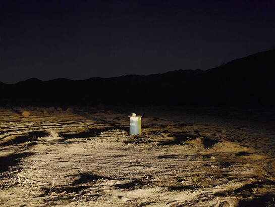 A color photograph of a white water storage barrel in a desert landscape, with mountains in the distance, at night.