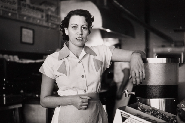 A black-and-white photograph of a White woman in a waitress uniform standing behind a diner counter with an arm propped on drink dispenser.