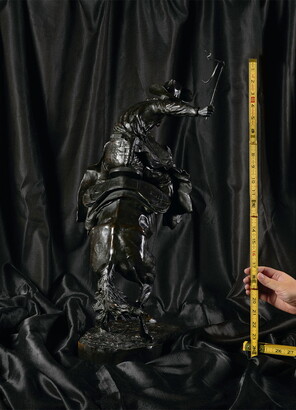 A photo of a hand holding measuring tape up to a bronze sculpture of a man on a bucking bronco.