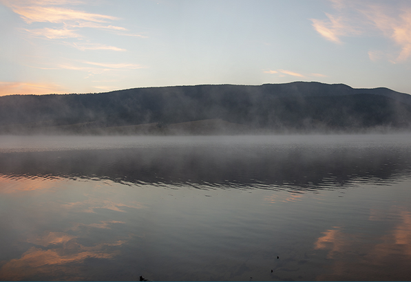 A detail of a color photograph of morning fog over a body of water with dark hills in the background.