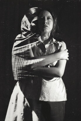 A black-and-white double-exposed photograph of a woman with her arms crossed across her torso; in one half she wears a dress and shawl and in the other a white shirt and pants.