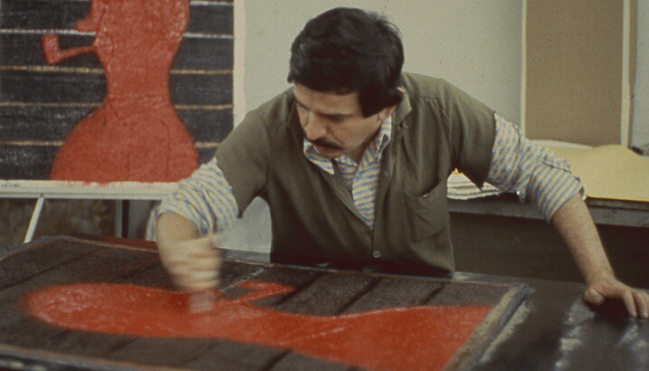 A man with dark hair and a mustache leans over a table adding red ink to a printing plate.